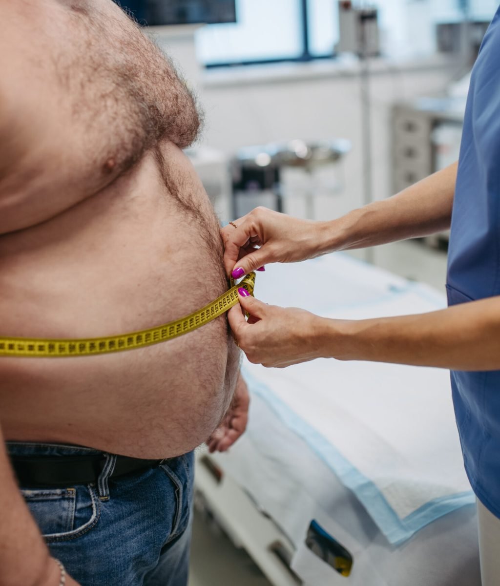 Female doctor measuring waist of overweight patient using a tape measure. Obesity affecting middle-aged men's health. Concept of health risks of overwight and obesity.